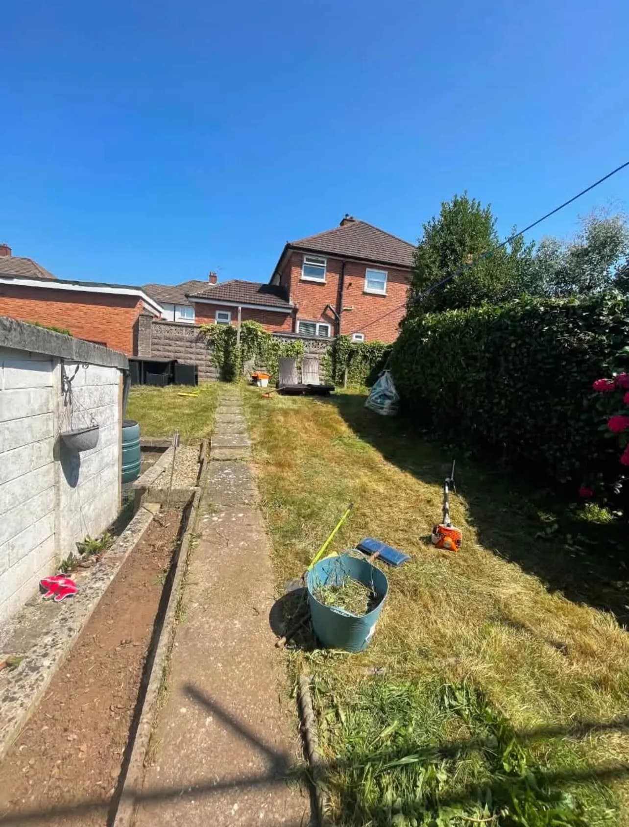 A long rear garden being tidied: a strimmer resting on the grass beside a tub of cuttings, with a concrete path running up the middle towards the house